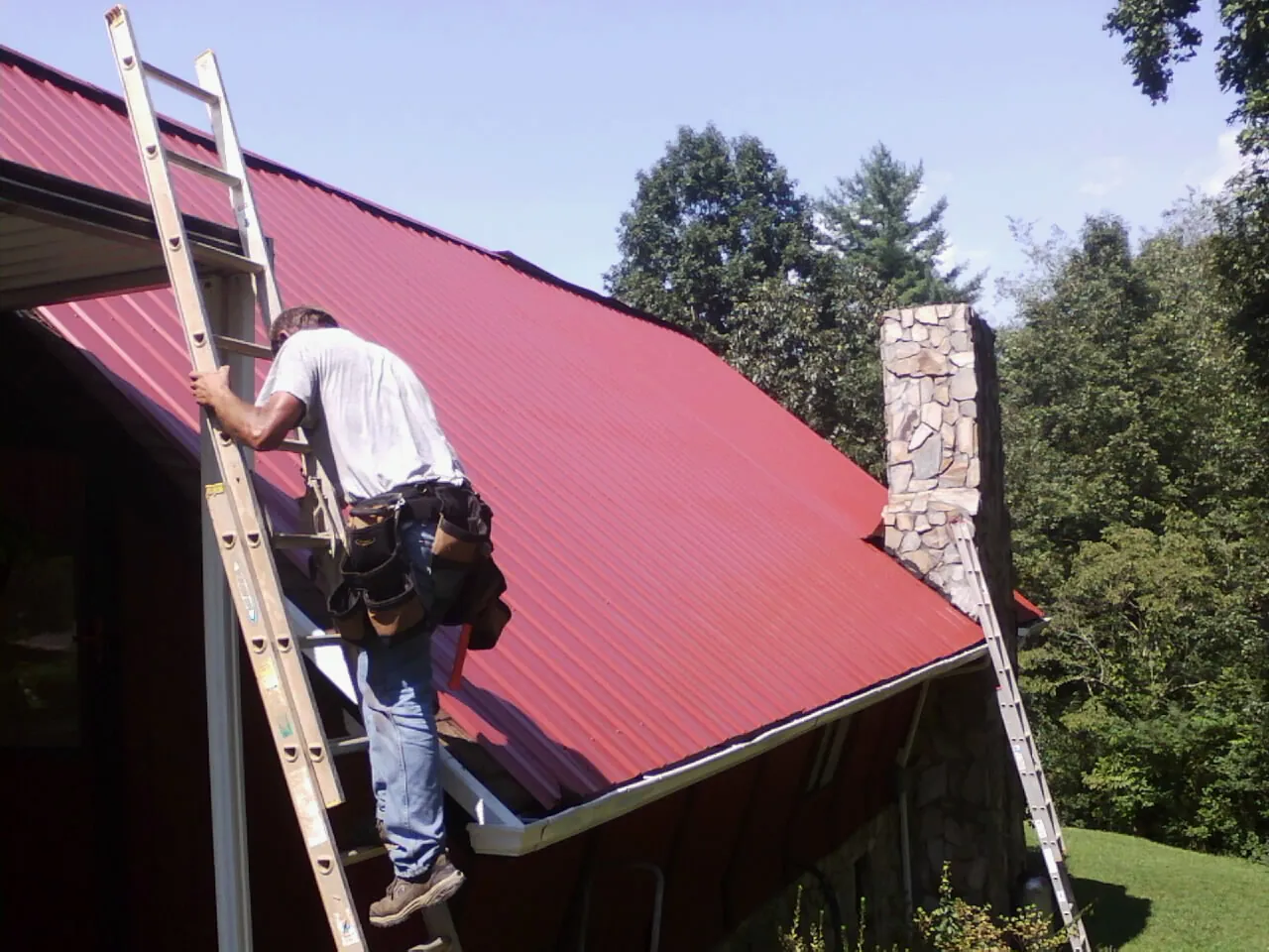 Roofer on a ladder beside a red metal roof and stone chimney — Soffit & Fascia Repair by Roofing Brothers in Lucasville